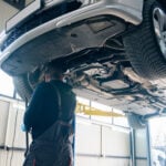 Serviceman checks drive train on a column lift in car service.